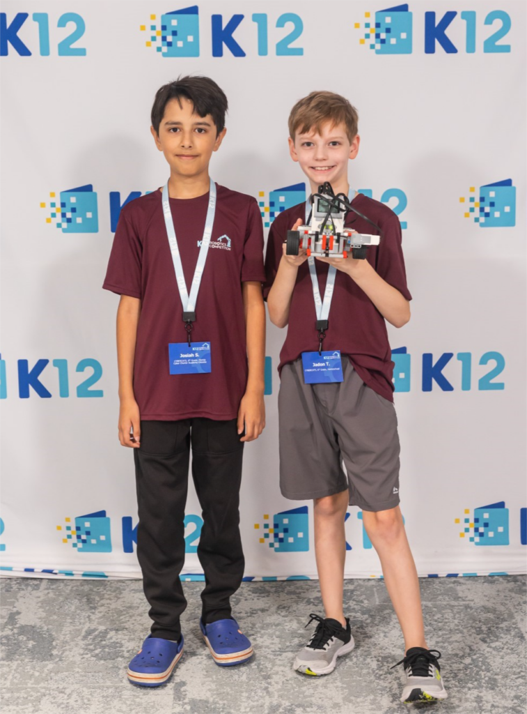Two boys in maroon shirts and name tags stand smiling in front of a K12 banner. One boy holds a robot model, symbolizing teamwork and innovation.