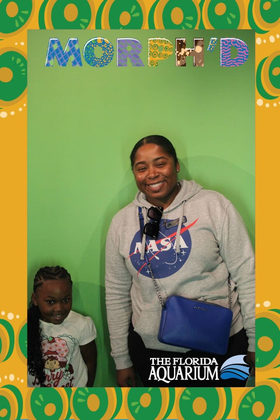 A smiling adult and child stand against a green background.