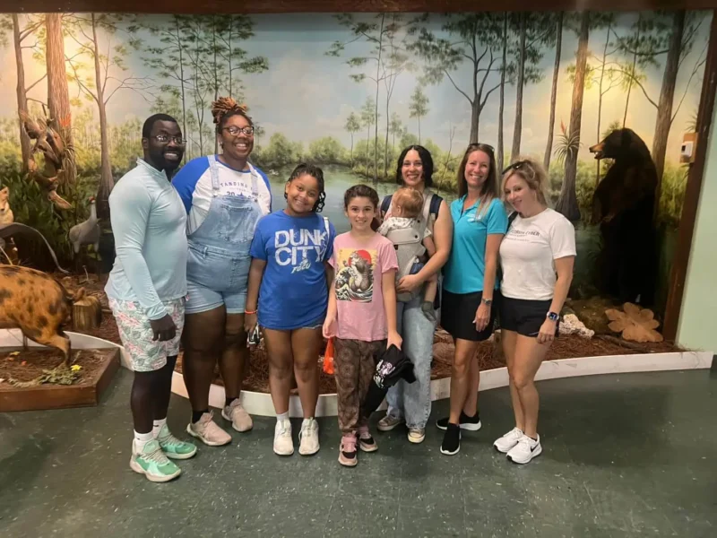 Seven people, including two children, stand smiling indoors against a forest-themed mural with animal figures.