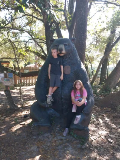 Two children sit on the lap of a large bear statue in a wooded park