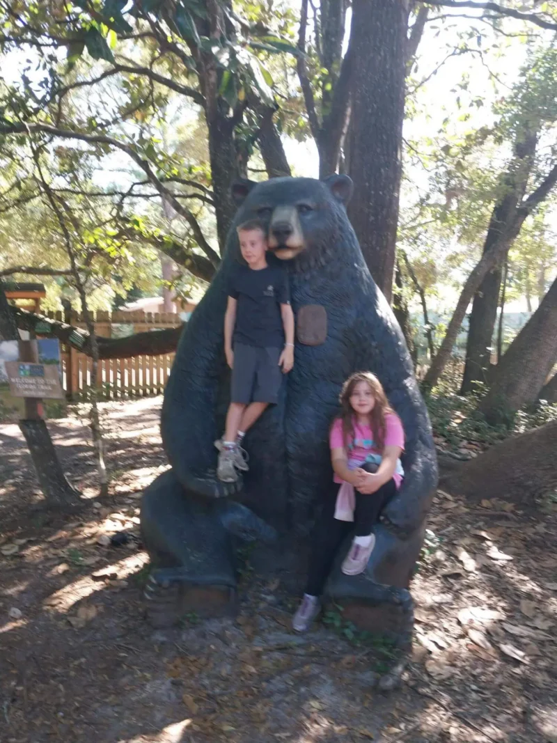 Two children sit on the lap of a large bear statue in a wooded park