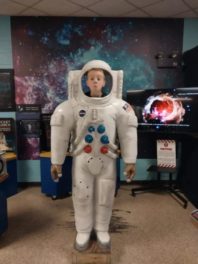 A young person stands behind a large astronaut cutout, appearing as if in a space suit. The room has cosmic-themed walls and displays, with a playful, educational tone.