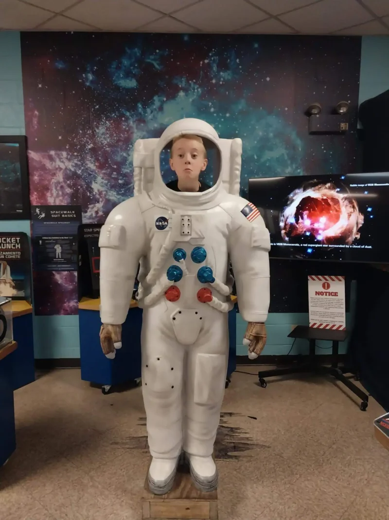 A young person stands behind a large astronaut cutout, appearing as if in a space suit. The room has cosmic-themed walls and displays, with a playful, educational tone.
