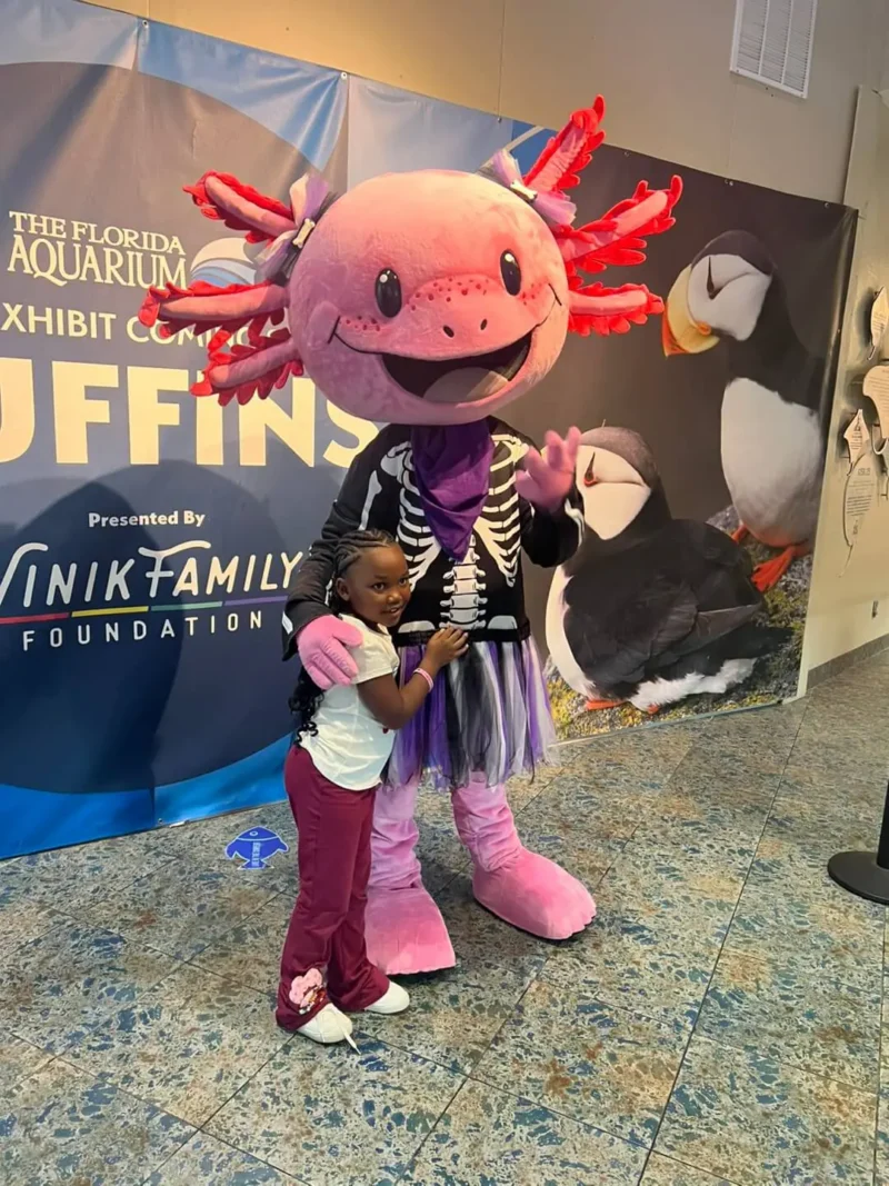 A child smiles and hugs a person in a pink axolotl costume at the Florida Aquarium.