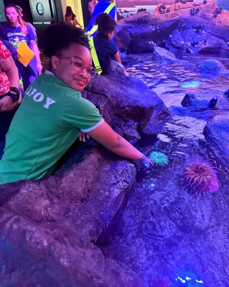 Person wearing glasses and a green shirt leans over a tank, interacting with sea anemones under blue lighting at an aquarium.
