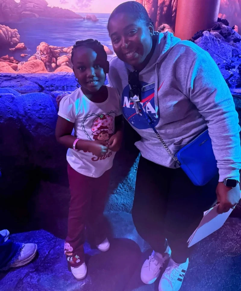 A smiling woman and a child stand close together in a brightly lit, colorful environment resembling an aquarium.