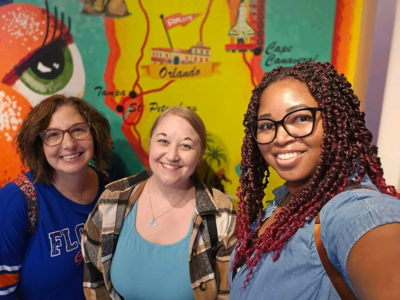 Three women smiling in front of a colorful map mural featuring Florida landmarks.