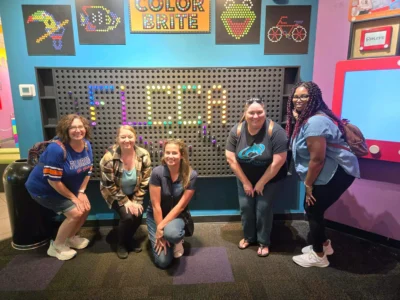 Five people pose smiling in front of a colorful interactive display with "FLCCA" written in lights.